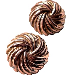 May include: Two copper-colored, spiral-design earrings. Each earring has a circular shape with a raised, swirling pattern. The earrings are set against a white background, highlighting their metallic sheen.