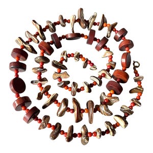 Puede incluir: Collar artesanal en espiral compuesto por cuentas de color marrón oscuro, marrón rojizo y beige. Presenta cuentas de diferentes formas y tamaños, unidas por pequeñas cuentas rojas y naranjas.