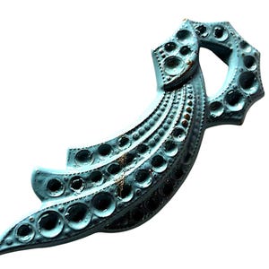 May include: A decorative, abstract metal object in a light blue hue with a textured surface. The design features a series of circular indentations and a curved, layered shape. The object has a distressed, aged appearance with hints of brown.
