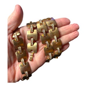 May include: Gold-toned necklace with a series of square links, each adorned with a row of small, clear crystals. The necklace is held in a hand, showcasing its design and sparkle. A vintage jewelry piece.