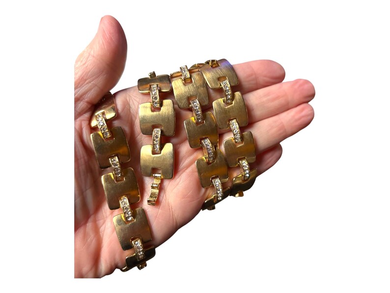 May include: Gold-toned necklace with a series of square links, each adorned with a row of small, clear crystals. The necklace is held in a hand, showcasing its design and sparkle. A vintage jewelry piece.