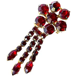 May include: A vintage gold-toned brooch featuring clusters of round, deep red rhinestones and three dangling strands of smaller, rectangular red stones. The brooch has a classic, elegant design.