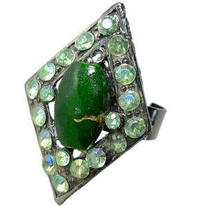 May include: A diamond-shaped ring featuring a dark metallic frame encrusted with small, light green circular rhinestones. A single, smooth, oval-shaped dark green bead with a small gold-coloured crack detail is set in the centre of the frame.