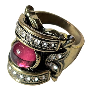 May include: A gold-toned metal ring featuring a large, round, deep pink cabochon stone set in the center. The band and setting are embellished with small, clear, sparkling stones arranged in rows and decorative patterns around the central gem.