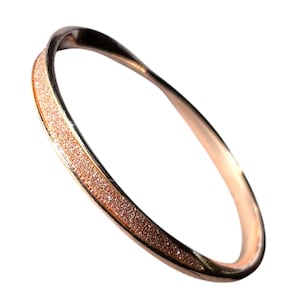 Può includere: Un bracciale a cerchio in oro rosa con una fascia strutturata e glitterata. Il bracciale presenta una finitura liscia e lucida sui bordi esterni e una fascia centrale scintillante. Il design è un anello continuo, che mostra uno stile moderno ed elegante.