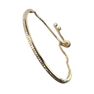 May include: A gold-coloured bracelet with a curved design, set with small, sparkling stones along the front. The bracelet has an adjustable chain and clasp. The jewellery is displayed against a white background.