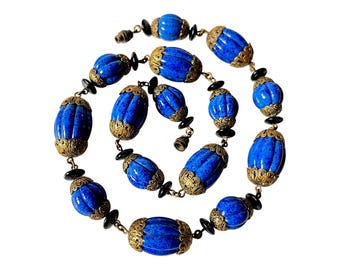 May include: A necklace featuring large, oval, cobalt blue beads with gold-colored filigree caps. The beads are interspersed with smaller black beads and connected by gold-toned chain links. The necklace is set against a white background.