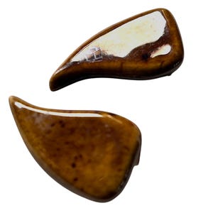 May include: Two glossy, brown ceramic pieces with a curved, leaf-like shape. The pieces have a speckled appearance and a high-shine finish, reflecting light. The design is organic and abstract, suitable for decorative purposes.