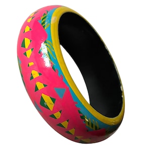 Puede incluir: Un brazalete de madera pintado con colores vibrantes. El brazalete presenta una base rosa brillante con patrones geométricos amarillos, verdes y azules. El interior es negro y los bordes están delineados en amarillo y azul. Circunferencia de unos 20 cm.