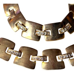 May include: Close-up of a gold-toned necklace and bracelet. The jewelry features square links with a brushed metal finish. Each link is connected by a smaller bar adorned with small, clear crystals, adding a touch of sparkle to the design.