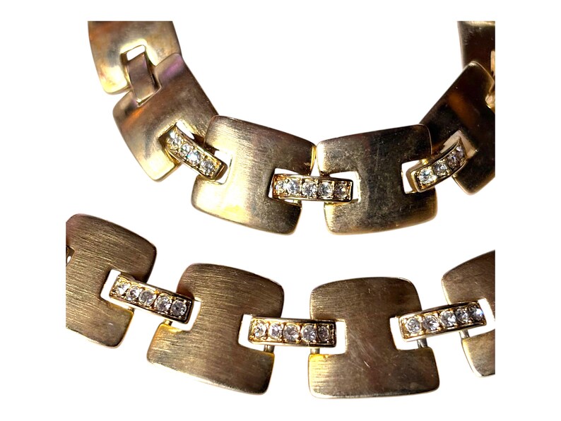 May include: Close-up of a gold-toned necklace and bracelet. The jewelry features square links with a brushed metal finish. Each link is connected by a smaller bar adorned with small, clear crystals, adding a touch of sparkle to the design.
