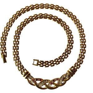Pode incluir: Colar dourado com um design de corrente tecida. O colar apresenta uma seção decorativa com pedras preciosas claras e vermelhas. O fecho é visível e o colar está sobre um fundo branco.