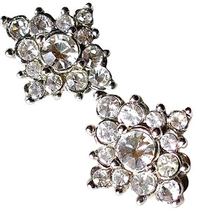 May include: A pair of silver-toned stud earrings, each featuring a square-shaped cluster of clear, faceted round rhinestones. The design centres on a larger circular stone surrounded by smaller stones, creating a multi-layered, reflective surface.