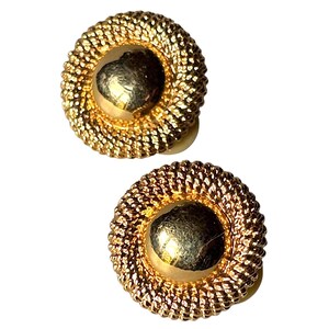May include: A pair of gold-toned clip-on earrings. Each earring features a textured, circular design with a central, smooth, gold-colored dome. The outer ring has a woven rope-like texture.