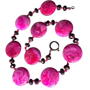 May include: A necklace with large, oval, pink agate stones and smaller, dark purple beads. The necklace has a silver clasp and is set against a white background.