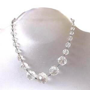 May include: A clear crystal bead necklace with varying sizes of faceted beads. The beads are strung on a silver-toned chain, creating a sparkling and elegant accessory. The necklace is displayed on a white mannequin, highlighting its design.