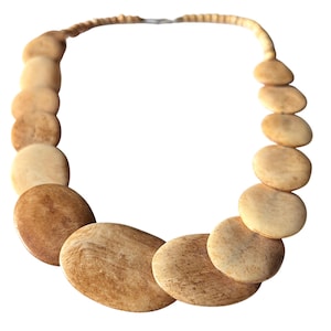 Puede incluir: Un collar beige con grandes cuentas ovaladas de aspecto natural y texturizado. Las cuentas están conectadas por un hilo más pequeño y redondeado, creando una pieza de joyería única y terrosa. El collar se presenta sobre un fondo blanco.