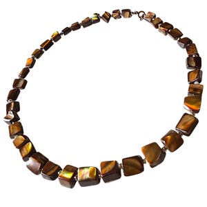 Puede incluir: Un collar compuesto por numerosas cuentas irregulares de forma cuadrada en tonos marrón oscuro, ámbar y dorado. Las cuentas presentan un acabado pulido e iridiscente con patrones de grano natural visibles, ensartadas para formar una cadena circular.