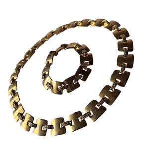 May include: A gold-toned necklace and bracelet set. The jewelry features a series of square-shaped links connected by smaller rectangular bars. Each square link is adorned with small, sparkling embellishments. The necklace and bracelet are displayed on a white background.