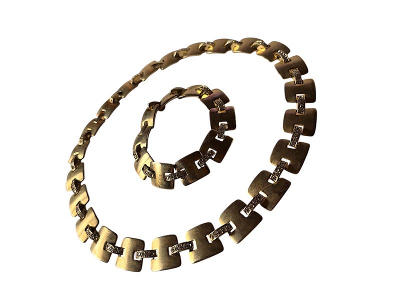 May include: A gold-toned necklace and bracelet set. The jewelry features a series of square-shaped links connected by smaller rectangular bars. Each square link is adorned with small, sparkling embellishments. The necklace and bracelet are displayed on a white background.