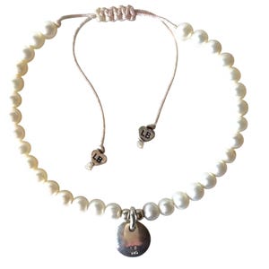 May include: A delicate pearl bracelet with a silver charm. The bracelet features a series of white pearls strung together with a light-colored cord. The charm is a small, round silver disc with the initials "LB" engraved on it.