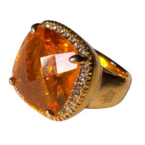 Puede incluir: Un anillo dorado con una gran gema naranja facetada. La gema está rodeada por una fila de pequeñas piedras transparentes. El anillo tiene una banda gruesa y un diseño decorativo en el lateral.