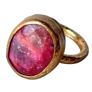 May include: A gold ring with a large, oval-cut gemstone in shades of pink and red. The ring's band has a textured, hammered finish. The gemstone is approximately 2 cm in length.