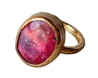 May include: A gold-toned ring featuring a large, faceted, oval-shaped gemstone with shades of pink and red. The ring's band has a textured, hammered appearance, adding to its unique design.