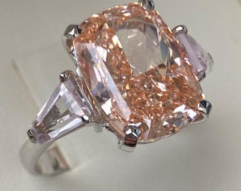 May include: A large, peach-colored gemstone ring with a rectangular center stone and two smaller side stones. The ring is set in a silver band and is displayed in a white box.