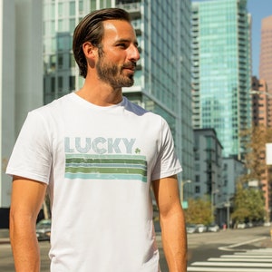 May include: White t-shirt with a retro-style graphic design featuring the word "LUCKY" in blue and green stripes with a green shamrock.