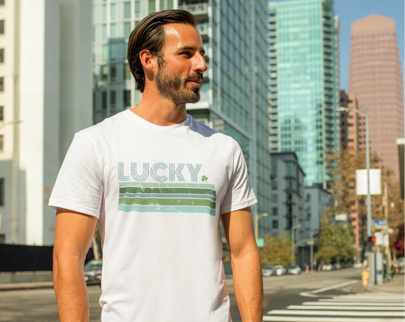 May include: White t-shirt with a retro-style graphic design featuring the word "LUCKY" in blue and green stripes with a green shamrock.
