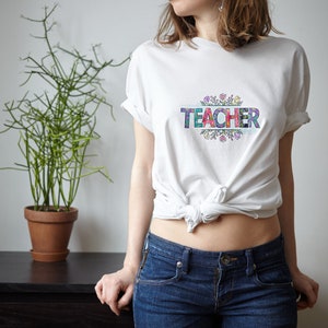 K&ouml;nnte beinhalten: Wei&szlig;es T-Shirt mit dem Wort "TEACHER" in bunten Buchstaben, umgeben von einem floralen Design.