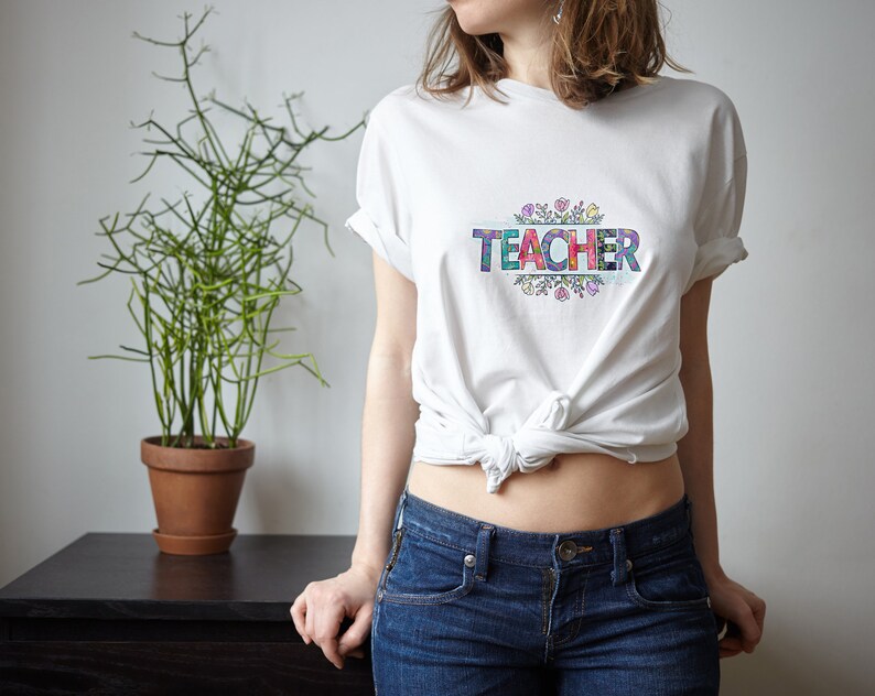 K&ouml;nnte beinhalten: Wei&szlig;es T-Shirt mit dem Wort "TEACHER" in bunten Buchstaben, umgeben von einem floralen Design.