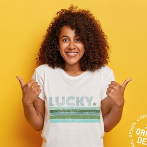 May include: A woman with curly brown hair is wearing a white t-shirt with a blue, green, and white striped design that says "LUCKY" with a shamrock. She is giving a thumbs up with both hands and smiling. The background is yellow.