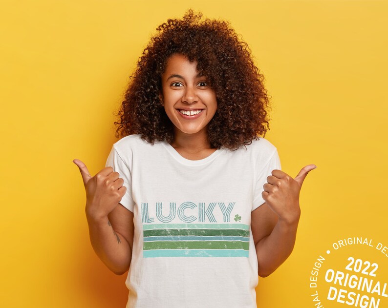 May include: A woman with curly brown hair is wearing a white t-shirt with a blue, green, and white striped design that says "LUCKY" with a shamrock. She is giving a thumbs up with both hands and smiling. The background is yellow.