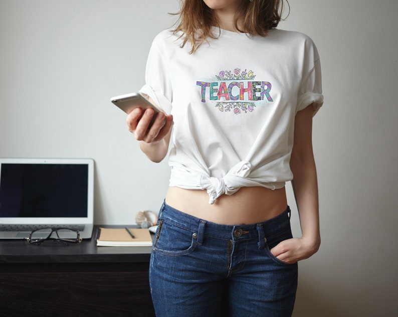 K&ouml;nnte beinhalten: Ein wei&szlig;es T-Shirt mit dem Wort "TEACHER" in bunten Buchstaben, umgeben von einem floralen Design. Das Shirt ist in der Taille gebunden und die Person tr&auml;gt eine blaue Jeans.
