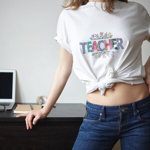 K&ouml;nnte beinhalten: Ein wei&szlig;es T-Shirt mit dem Wort "TEACHER" in bunten Buchstaben, umgeben von einem floralen Design.