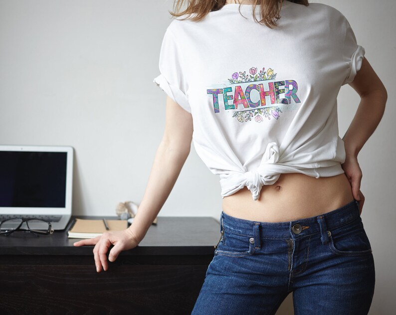 K&ouml;nnte beinhalten: Ein wei&szlig;es T-Shirt mit dem Wort "TEACHER" in bunten Buchstaben, umgeben von einem floralen Design.