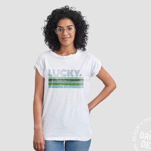 May include: A white t-shirt with a green and blue striped design and the word "LUCKY" in a retro font. A small shamrock is next to the word "LUCKY".