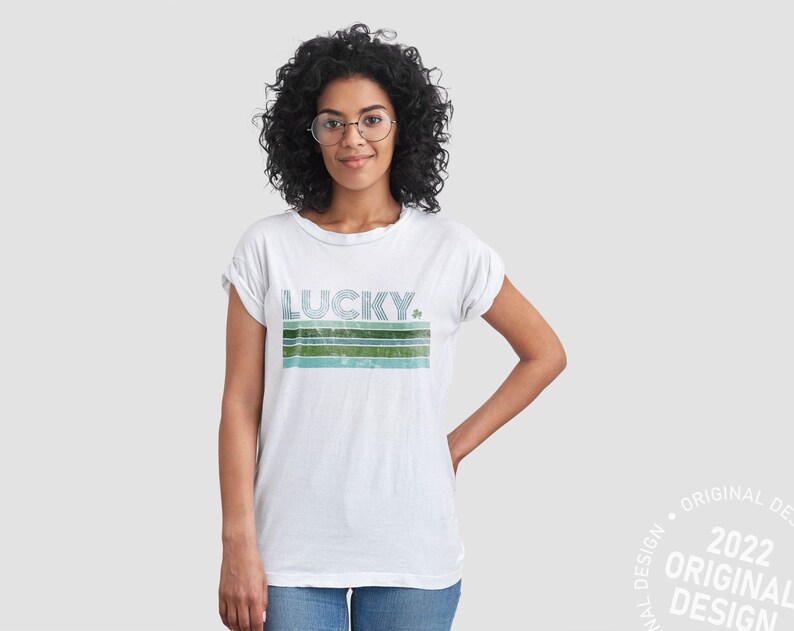 May include: A white t-shirt with a green and blue striped design and the word "LUCKY" in a retro font. A small shamrock is next to the word "LUCKY".