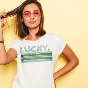 May include: White t-shirt with a retro-style graphic that says "LUCKY" with a green shamrock. The graphic is made up of blue, green, and white stripes.