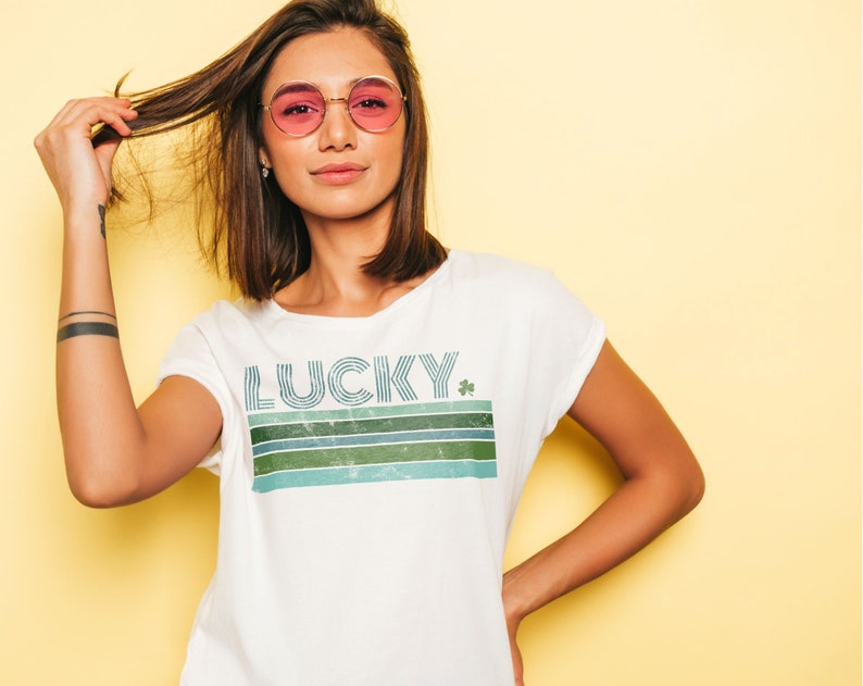 May include: White t-shirt with a retro-style graphic that says "LUCKY" with a green shamrock. The graphic is made up of blue, green, and white stripes.