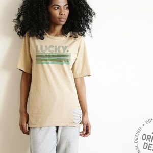 May include: A beige t-shirt with a distressed look featuring a retro-style graphic that says "LUCKY" with a shamrock. The graphic is in shades of green and blue.