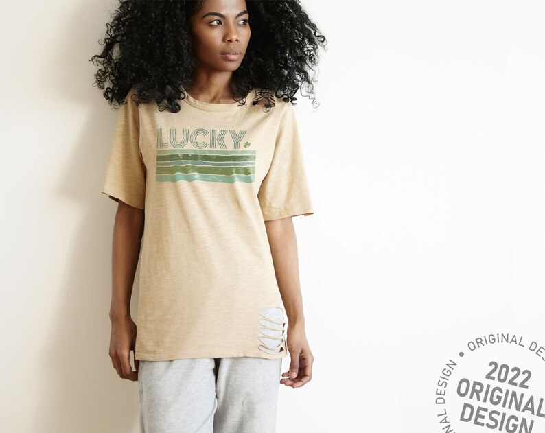 May include: A beige t-shirt with a distressed look featuring a retro-style graphic that says "LUCKY" with a shamrock. The graphic is in shades of green and blue.