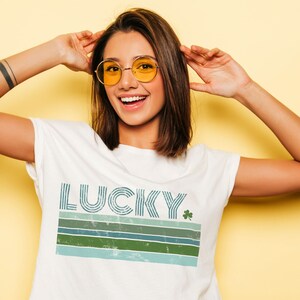 May include: White t-shirt with a retro-style graphic that says "LUCKY" with a green shamrock. The graphic features blue and green stripes.