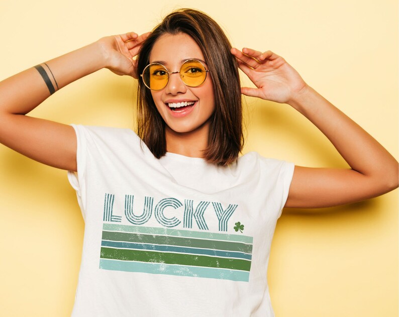 May include: White t-shirt with a retro-style graphic that says "LUCKY" with a green shamrock. The graphic features blue and green stripes.
