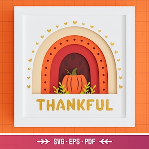 May include: A white frame with a layered paper craft design of a rainbow with a pumpkin in the center. The rainbow is made of orange, red, and cream paper. The pumpkin is orange with a green stem. The words "THANKFUL" are written in gold glitter letters at the bottom of the frame.