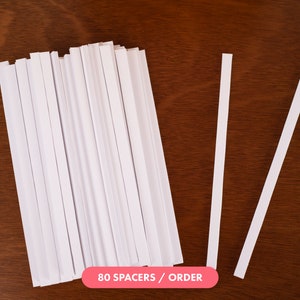 May include: A stack of 80 white paper spacers, with two individual spacers lying on a brown wooden surface. The text "80 SPACERS / ORDER" is printed on a pink rectangle below the stack of spacers.