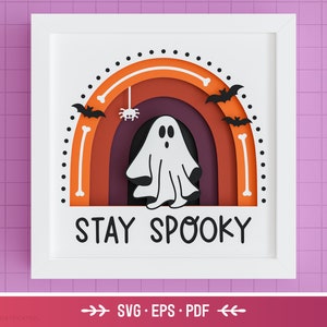 May include: A white frame with a white background and a rainbow layered paper craft design. The rainbow is orange, red, and brown. A white ghost is in the center of the rainbow. There are black bats and a spider on the rainbow. The text "STAY SPOOKY" is below the rainbow.