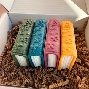 May include: Four small decorative book-shaped soaps in a box. The soaps are green, blue, pink, and yellow. The soaps have a decorative design on the front and are sitting on brown crinkle paper.